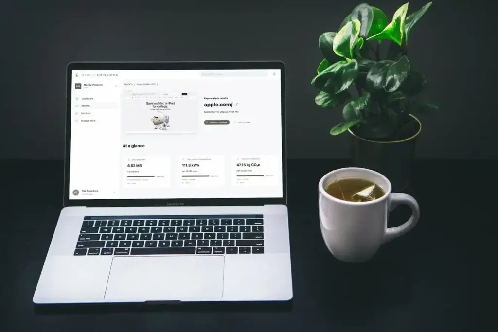 A MacBook Pro that displays the Merely Emissions platform with a successful scan of Apple.com. On the right side of the laptop there is a small plant in a pot, and a cup of tee.